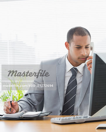 Concentrated young businessman using computer and phone at office desk