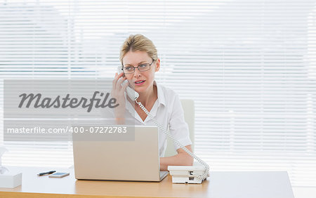 Concentrated young businesswoman using laptop and phone at office desk