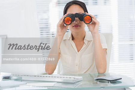 Serious businesswoman looking through binoculars at office desk