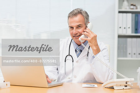 Smiling doctor using laptop and talking on phone at desk in his office at the hospital