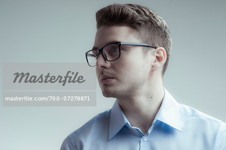 Close-up portrait of young man wearing eyeglasses and blue shirt, looking to the side, studio shot on white background