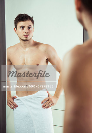 Portrait of young man with towel wrapped around waist, looking in bathroom mirror, studio shot