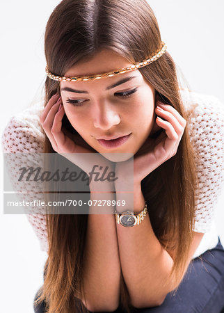 Portrait of young woman with long, brown hair, wearing headband, leaning on elbows and looking downwards, studio shot on white background