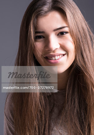 Close-up portrait of young woman with long brown hair, smiling and looking at camera, studio shot on grey background