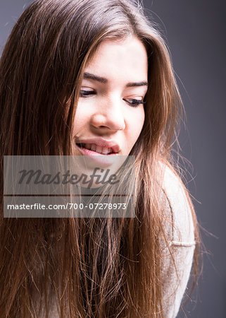 Close-up portrait of young woman with long brown hair, smiling and looking downwards, studio shot on grey background