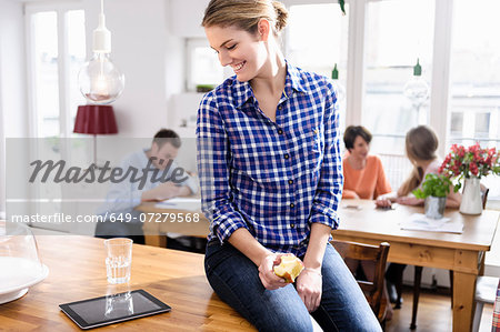 Young woman eating apple looking at tablet screen smiling, people in background