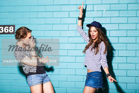 a girl takes picture of her friend in front of light blue brick wall