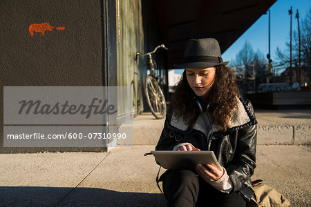 Teenage girl sitting on curb of sidewalk outdoors, wearing fedora and using tablet computer, Germany