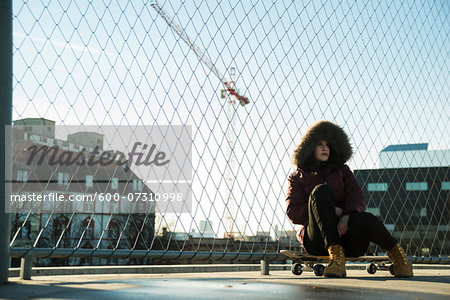 Teenage girl wearing winter coat, sitting on skateboard outdoors, next to chain link fence near comercial dock, Germany