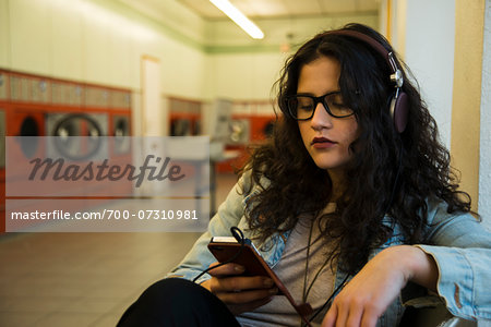 Teenage girl sitting in laundromat, wearing headphones and listening to music on smart phone, Germany