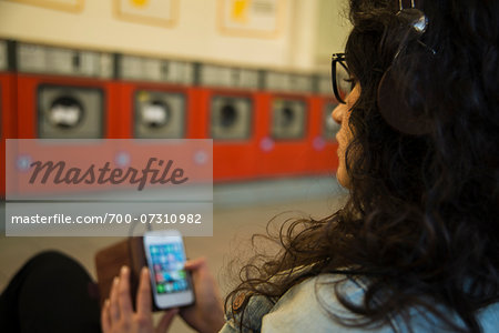 Teenage girl sitting in laundromat, wearing headphones and using smart phone, Germany