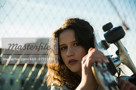 Close-up portrait of teenage girl standing outdoors next to chain link fence, holding skateboard and looking at camera, Germany