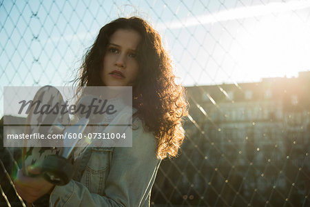 Portrait of teenage girl standing outdoors next to chain link fence, holding skateboard, Germany