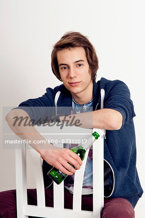 Portrait of teenage boy sitting on chair holding bottle of beer, smiling and looking at camera, studio shot on white background