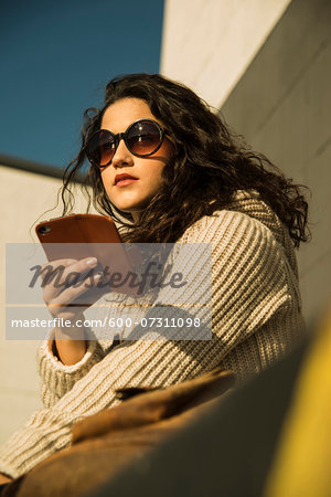 Close-up portrait of teenage girl outdoors wearing sunglasses, sitting next to building and holding smart phone, Germany