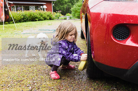 3 year old girl washing a red car while little white dog is watching, Sweden