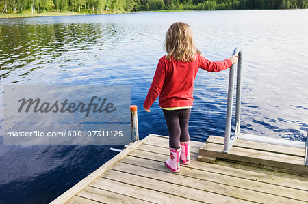 3 year old girl in red shirt on wooden dock looking at a lake, Sweden