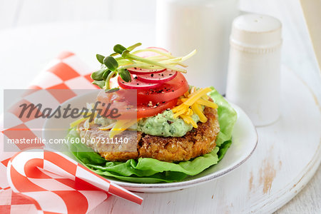 A gluten free chickpea burger with lettuce, chickpeas, guacamole, shredded cheddar cheese, tomatoes, red onion, and sprouts on a white plate with a diner style checkered paper and salt and pepper shakers.