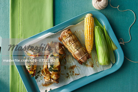 Chicken breast stuffed with red peppers, herbs and corn wrapped in a corn husk and cooked on the barbeque. Shown with a raw cob of corn and twine to tie the husks on a tabletop.