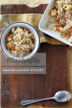 Turkey and white rice casserole with parsnips, carrots, and fresh parsley on a wooden cutting board on a table top. Whole ceramic casserole dish beside a single serving meal in a bowl and a serving spoon.