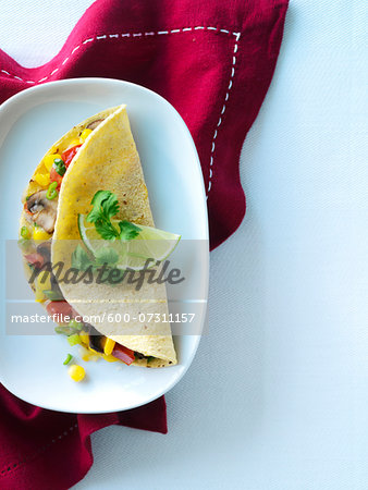 One vegetarian quesadilla in a yellow corn tortilla with cilantro, lime, mushrooms, corn, red pepper, melted cheese, and green onion. Mexican quesadilla is shown on a white cereamic plate with a red napkin on a tabletop.