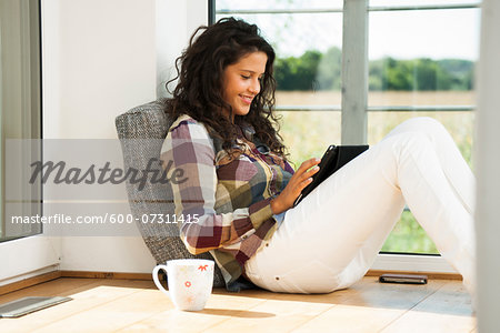 Teenage girl sitting on floor by window and reading on tablet computer, Germany