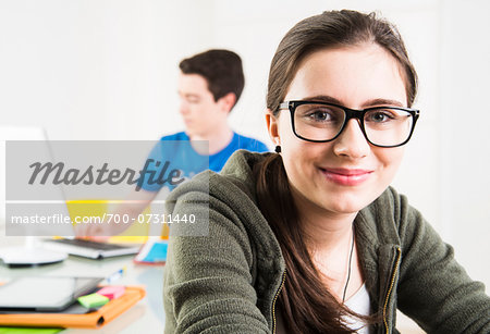 Teenage girl wearing eye glasses and looking at camera, with teenage boy working on project using computer in background, studio shot