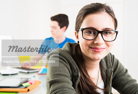 Teenage girl wearing eye glasses and looking at camera, with teenage boy working on project using computer in background, studio shot Teenage girl wearing eye glasses and looking at camera, with teenage boy working on project using computer in background, studio shot