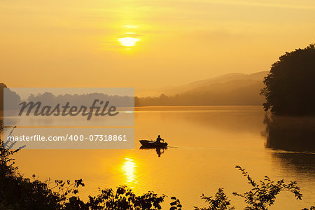 A lone fisherman moves out on a lake in a small boat as the sun breaks through dense morning fog.