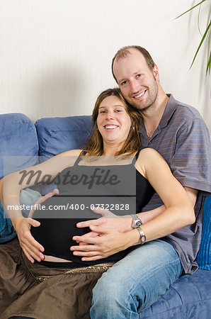 Smiling man embracing happy pregnant woman sitting on couch holding tummy