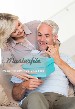 Portrait of a smiling woman surprising mature man with a gift on sofa at home