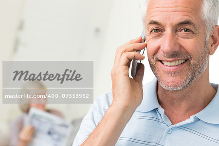 Closeup of a mature man using cellphone with woman reading newspaper in background at home