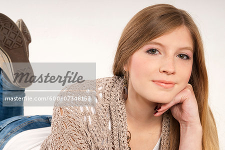 Close-up portrait of young, blond, long-haired woman lying on stomach, looking at camera and smiling, studio shot on white background