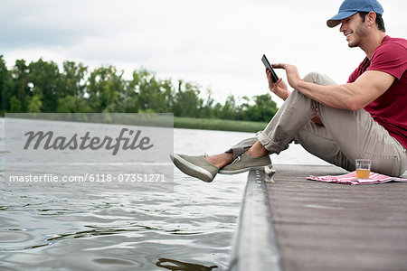 A man seated on a jetty by a lake, using a digital tablet.