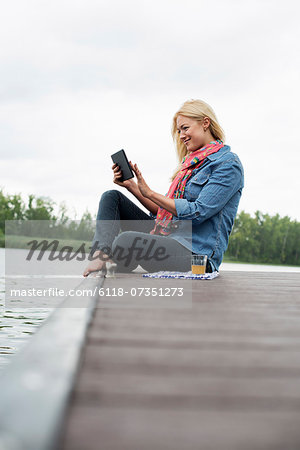 A woman sitting on a jetty by a lake, using a digital tablet.