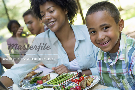 A family picnic meal in the shade of tall trees. Parents and children helping themselves to fresh fruits and vegetables.