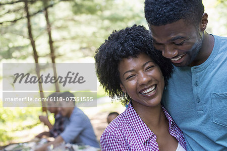 A family picnic meal in the shade of tall trees. A couple hugging each other.