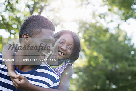 A man giving a child a piggyback, in the shade of trees on a summer day.