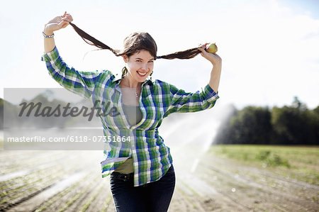 A girl in a green checked shirt with braids standing in a field with sprinklers working in the background.