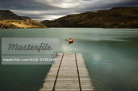 A young man in mid air, diving off a wooden pier, into calm lake surrounded by mountains in Torres del Paine National Park, Chile.