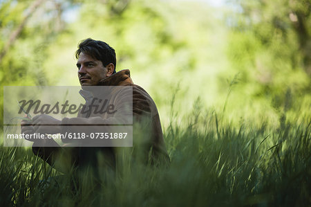 A man outdoors, sitting on the ground, looking thoughtful and contemplative.