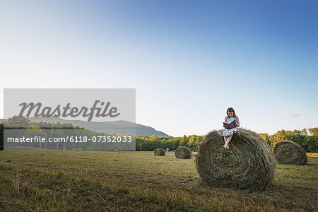 A field full of tall rounded hay bales, and a young girl sitting on the top of one large bale.