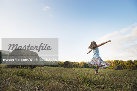 A field full of tall rounded hay bales, and a young girl dancing with her arms outstretched on the stubble field.