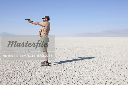 A man aiming a hand gun, holding it with his arm outstretched, standing in a vast, barren desert.