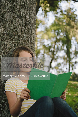 Nine year old girl sitting beneath tree, reading book