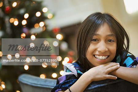 A young girl smiling and leaning over a chairback, in front of a Christmas tree.