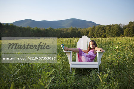 A young girl sitting in a traditional wooden Adirondack style chair in a field at evening light.
