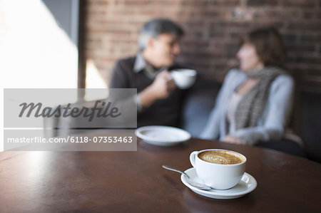 Two people sitting in a coffee shop. A man holding a white china of and drinking. Sitting beside a woman. A table with a large full cup of  cappuccino coffee.