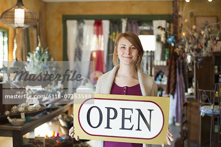 A woman standing in an antique store, holding an OPEN sign. Displays of goods all around her.