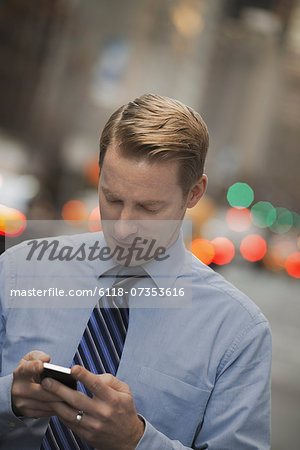 A man in a shirt and tie checking his cell phone, standing on a busy street.